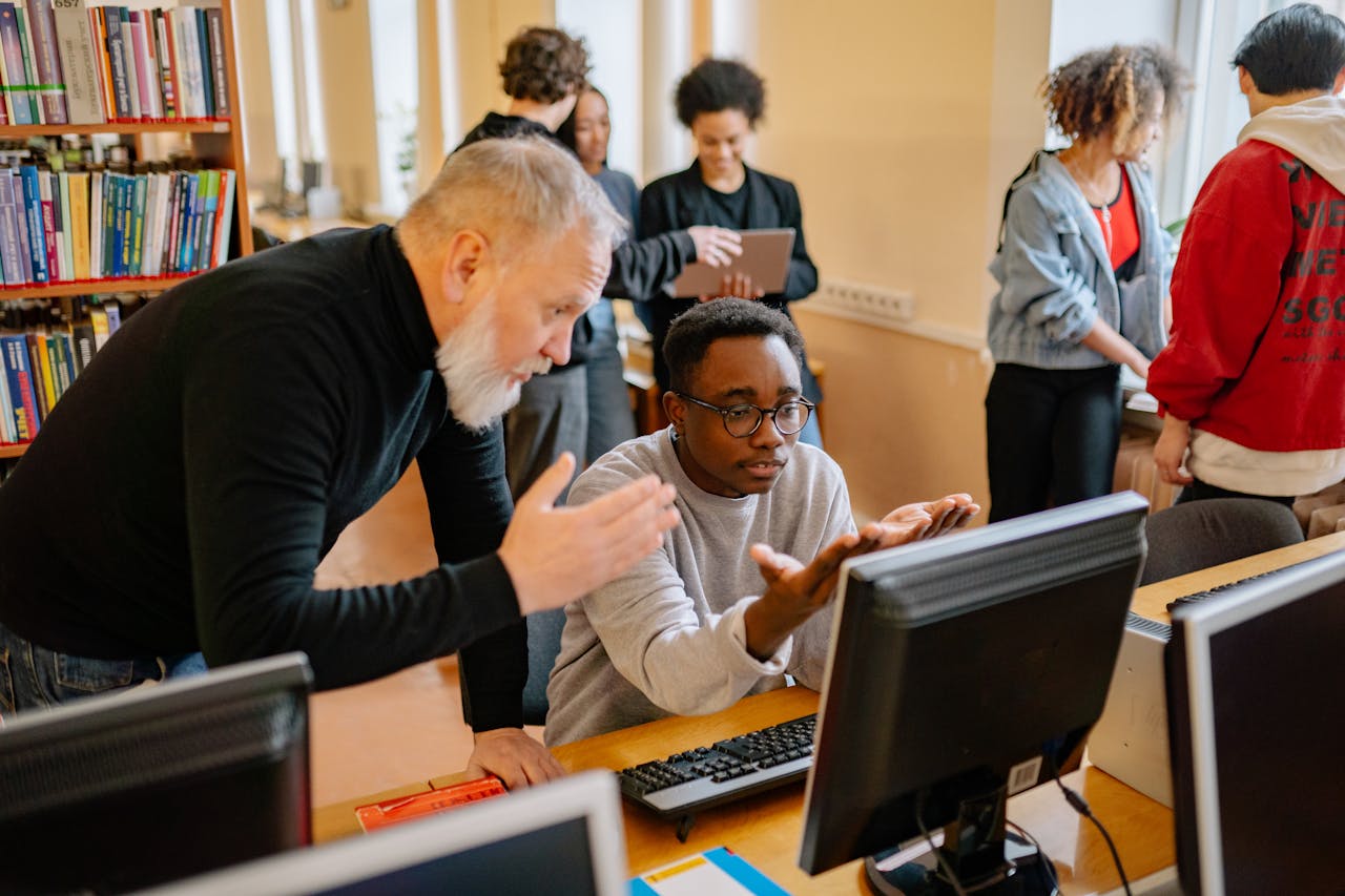 Teacher helping a student at a computer in a busy library setting, engaging in educational interaction.